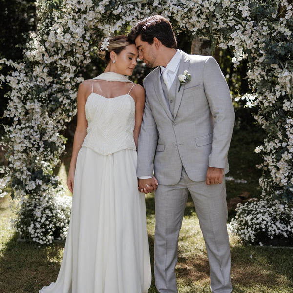Casal diante de parede de flores, noivo de terno cinza claro
